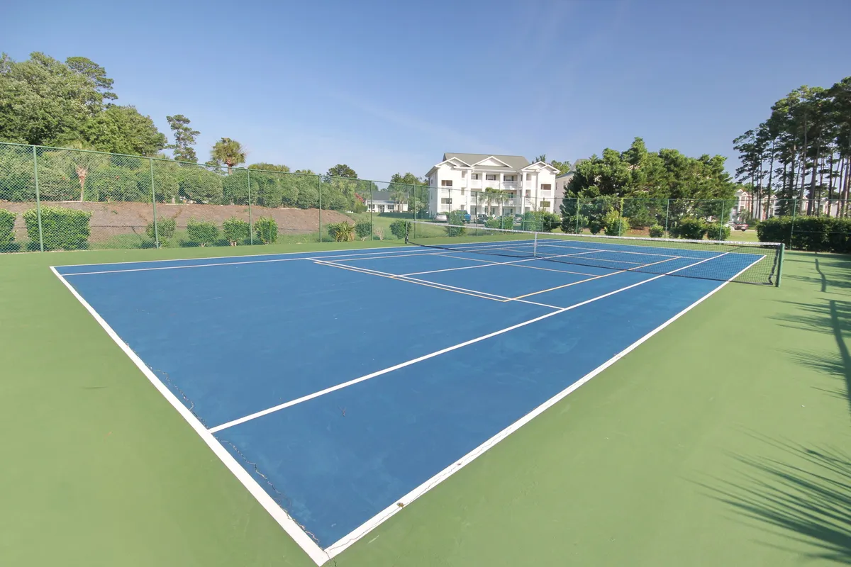 The photo captures a well-kept tennis court bordered by greenery, with a community building visible in the background. The vibrant court surface invites residents to engage in friendly matches or leisurely play, while the surrounding scenery contributes to a lively, active atmosphere.