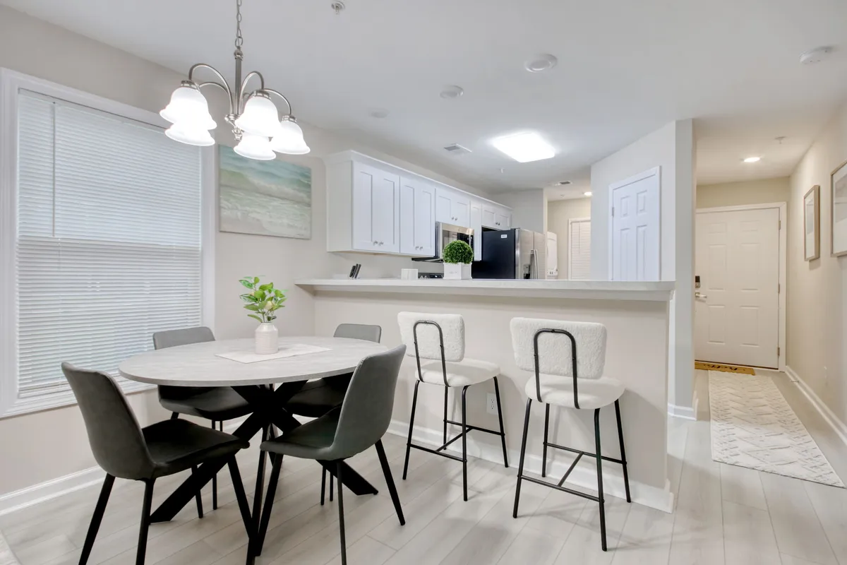 The open dining area features a welcoming dining table, elegant bar stools at a kitchen island, a stylish chandelier, and a touch of greenery from a potted plant. The setting is perfect for shared meals, morning coffee, or festive gatherings, all in a bright and airy environment.
