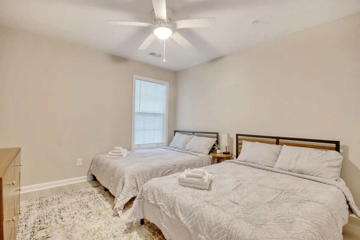 A spacious bedroom featuring two comfortable beds, a ceiling fan, and a bright window that brings in natural light. The room has a calming and welcoming atmosphere, ideal for families or friends sharing the space.