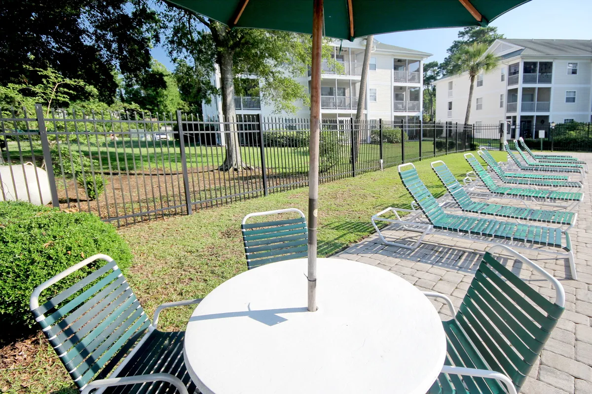 The image features a resort-style pool area with inviting poolside chairs, an umbrella providing shade, a table, and a landscaped garden. The setting is sunny and vibrant, suggesting a perfect place for families to relax and have fun together in the mornings.