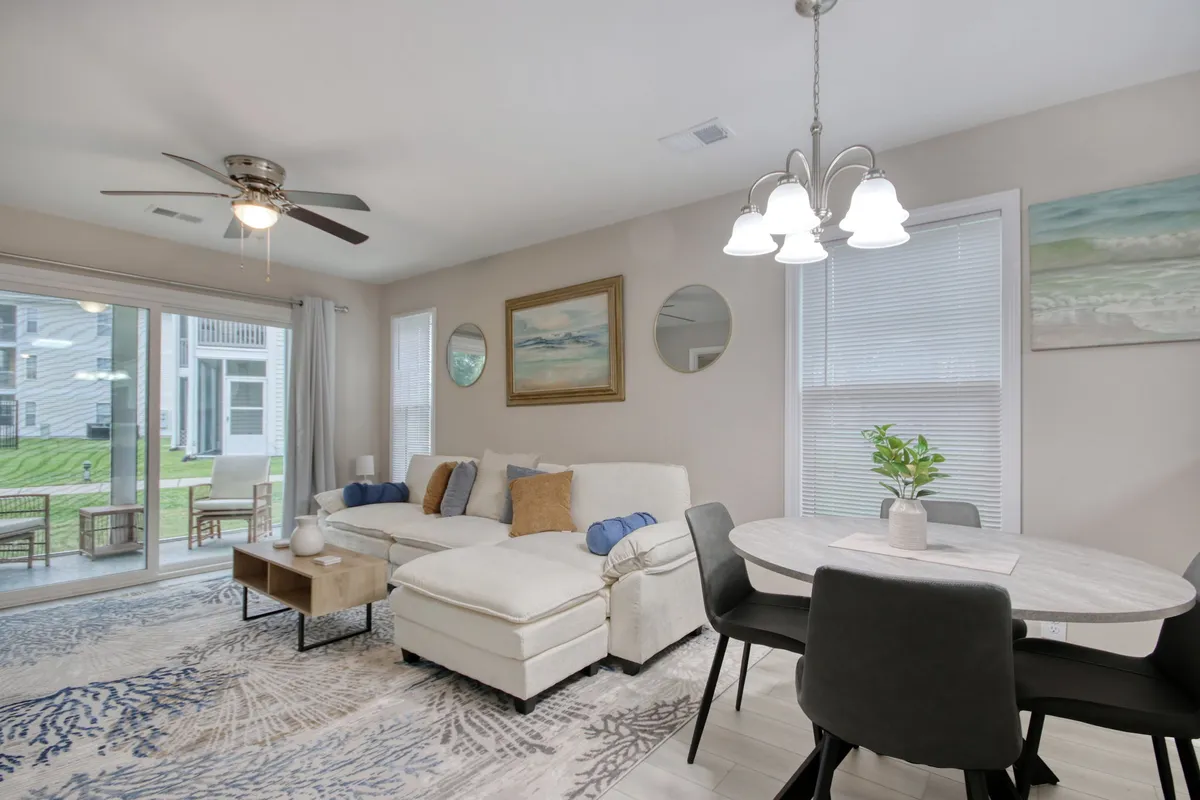 A welcoming open-concept living and dining area featuring a plush sofa, a spacious dining table, and vibrant artwork on the wall. Large windows allow sunlight to flood the space and offer expansive views of the green fairways outside, while the arrangement invites group gatherings, reading, or casual meals.