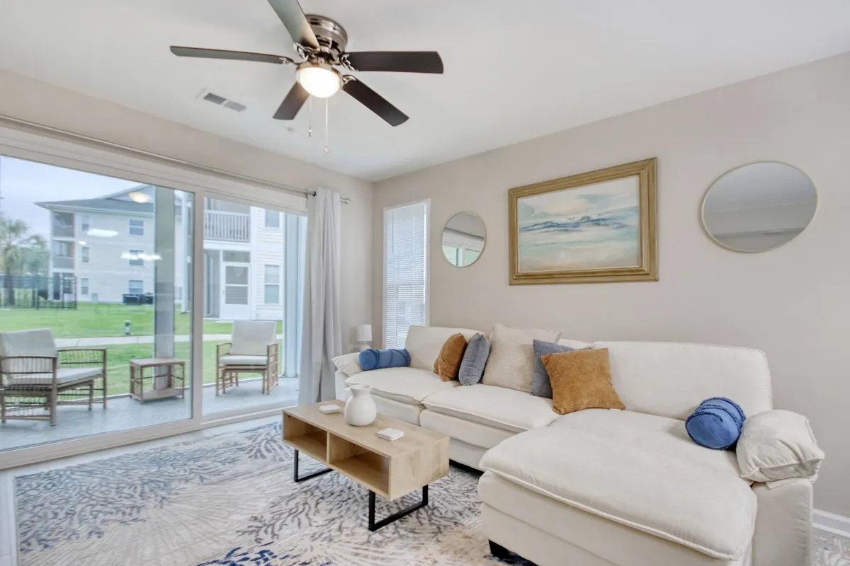 The image showcases a bright, inviting living room with a comfortable sofa, stylish coffee table, tasteful artwork, and mirrored accents. Sunlight filters in, casting a warm, welcoming glow—perfect for relaxing with friends or family after a day on the golf course.