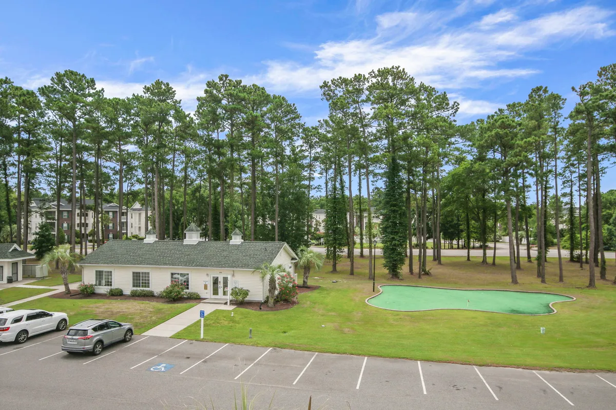 An exterior shot of a tranquil green pond encircled by leafy trees, with a community building and parking area nearby. The peaceful waters, gathering spaces, and natural surroundings invite relaxation, socialization, and nature walks.